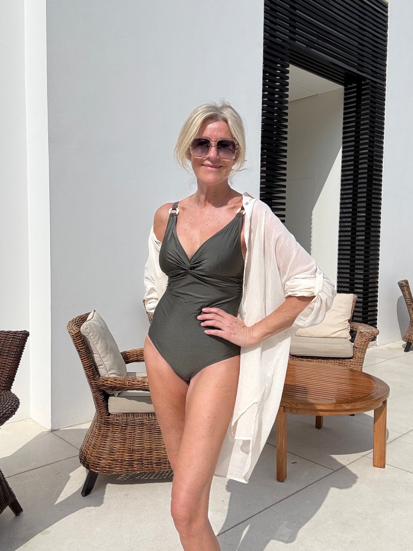 En kvinde i solbriller og badedragt har en Missya Siena Champagne-strandkjole på og står ved nogle kurvestole og et træbord.
