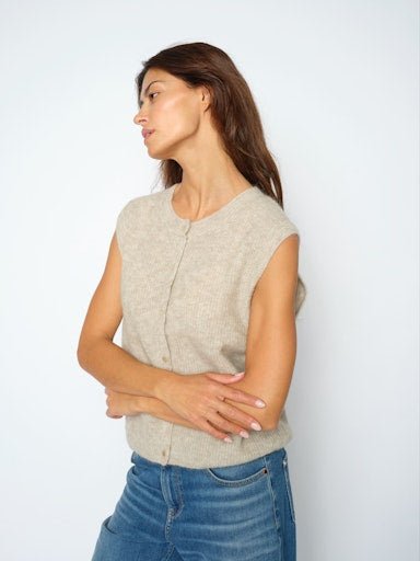En kvinde iført en MOS MOSH Almine Thora Sand-vest og blå jeans står med armene over kors og kigger mod venstre foran en ensfarvet baggrund.