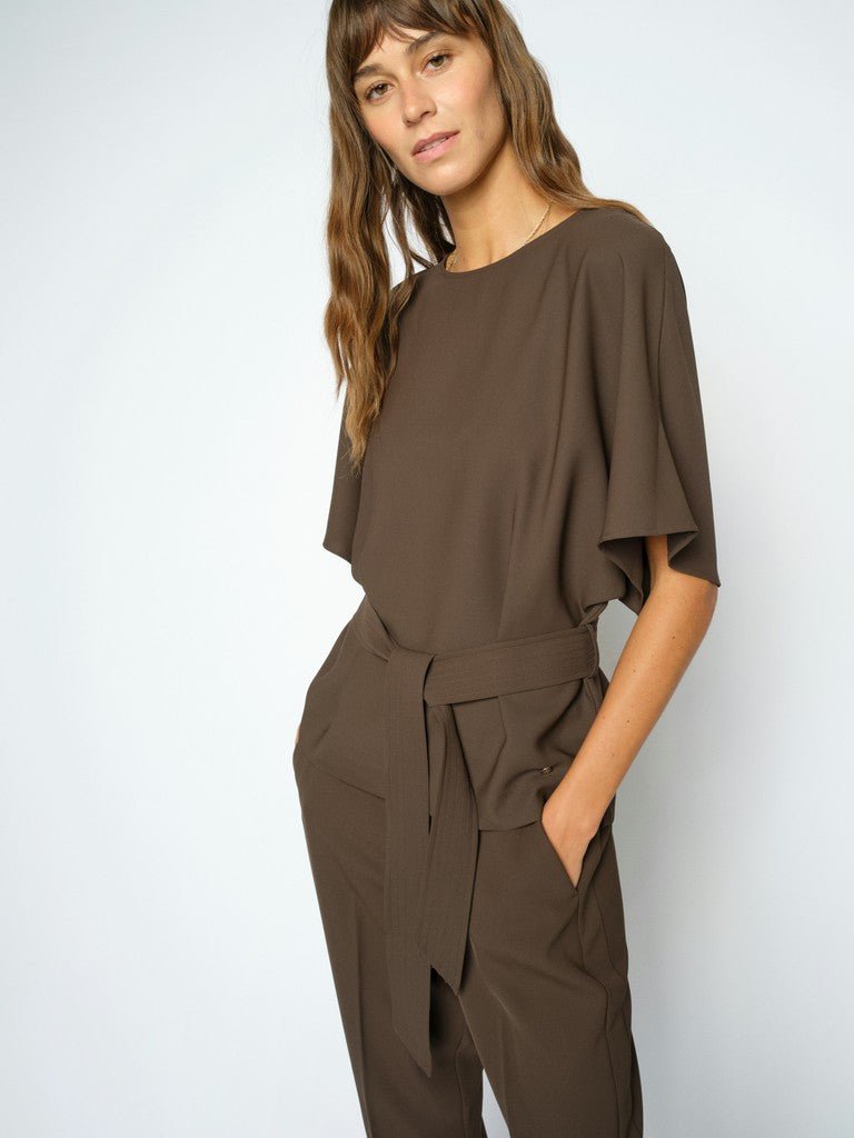 En kvinde iført en MOS MOSH Rikala Miley mørkebrun jumpsuit med bælte står foran en ensfarvet baggrund.