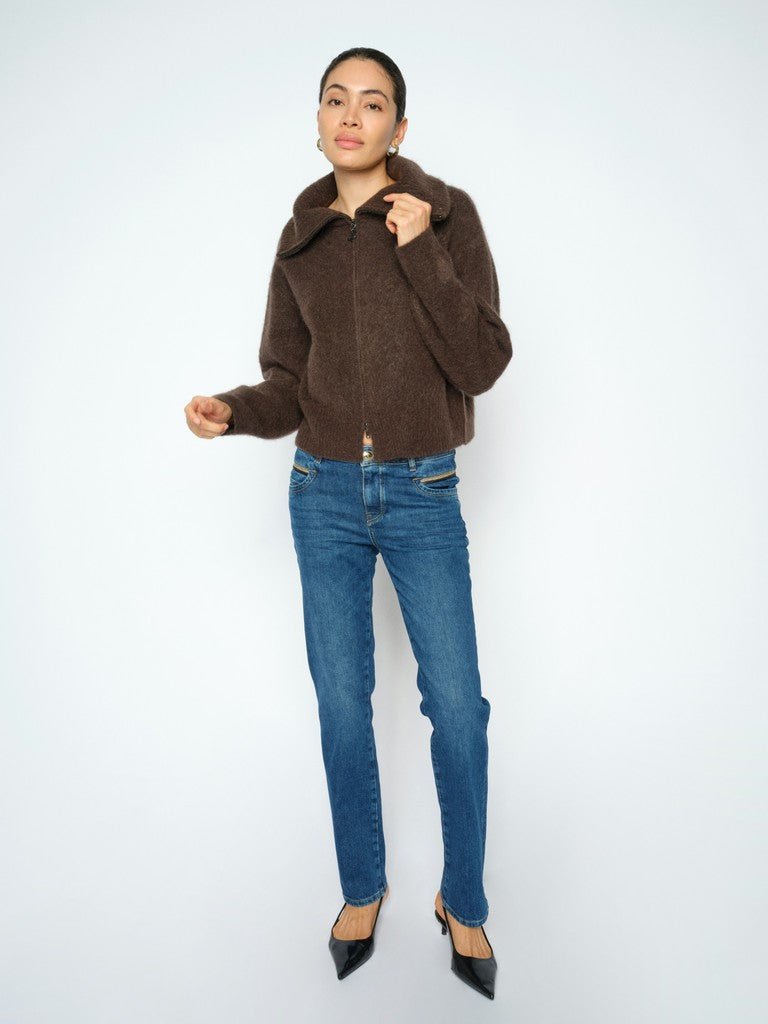 En kvinde poserer i en brun sweater med lynlås, MOS MOSH Carla Naomi L.32-jeans og sorte højhælede sko foran en hvid baggrund.