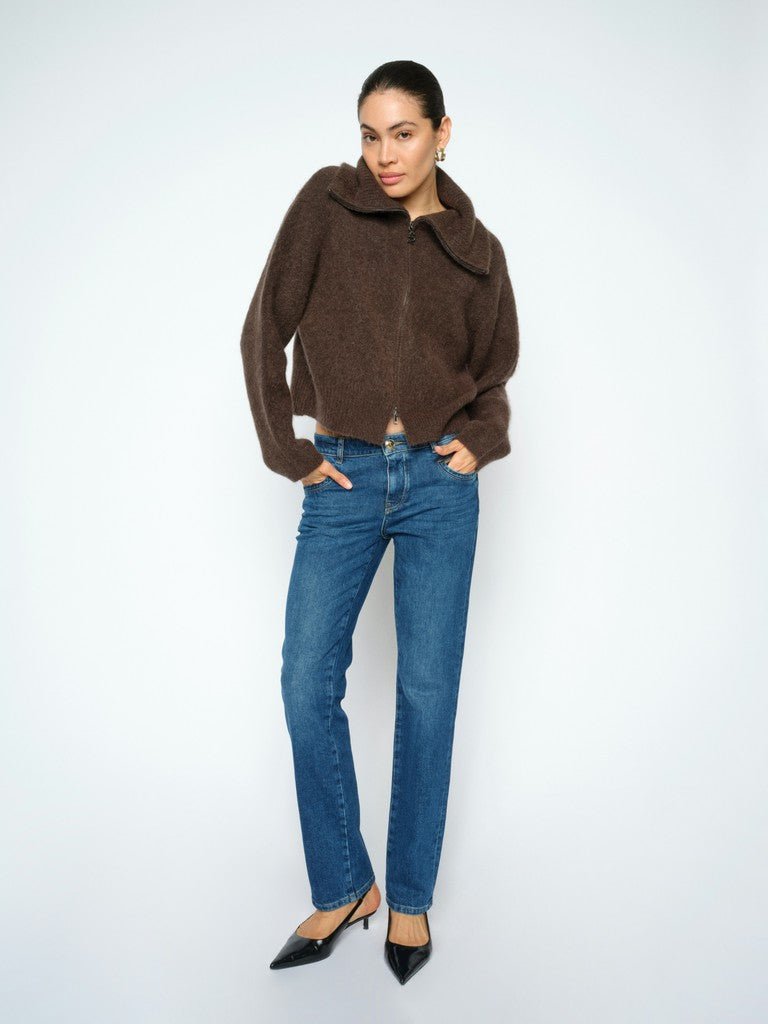 En kvinde iført en brun sweater med lynlås, MOS MOSH Carla Naomi l.32 denimjeans og sorte højhælede sko står foran en hvid baggrund.