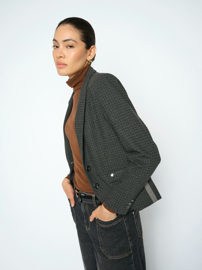 En person, der er klædt i en brun rullekravebluse, mørke jeans og en MOS MOSH Josa Ternet-blazer, står med siden til foran en lys baggrund.