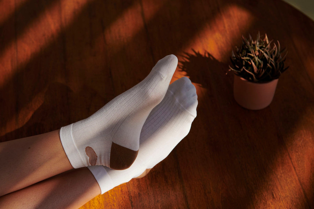 To par Minna Off White-sokker fra MP Denmark ligger på et stykke træ; den ene har et hul. En potteplante står ved siden af.