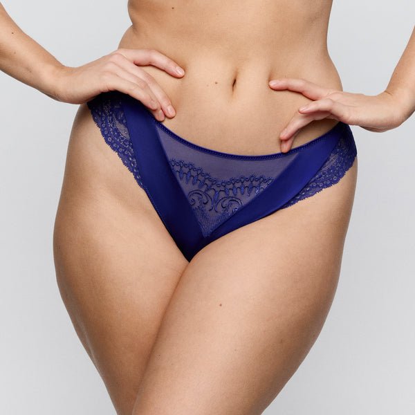 Model iført en blå PrimaDonna Shimla Crazy Blue G-streng, med hænderne på hofterne, fotograferet fra midten af overkroppen til lårene, på en ensfarvet baggrund.