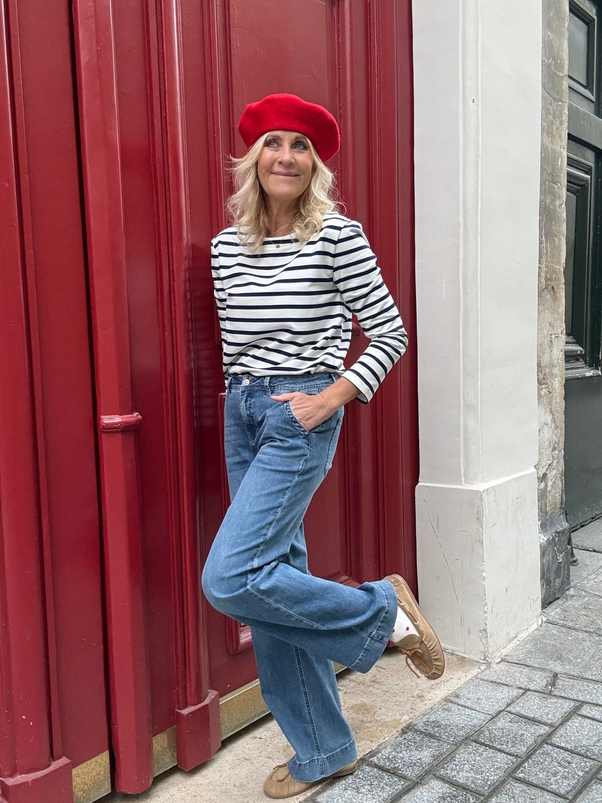 En kvinde med rød baret, stribet skjorte og et par Red Button Colette L.31-jeans står ved en rød dør i byen.