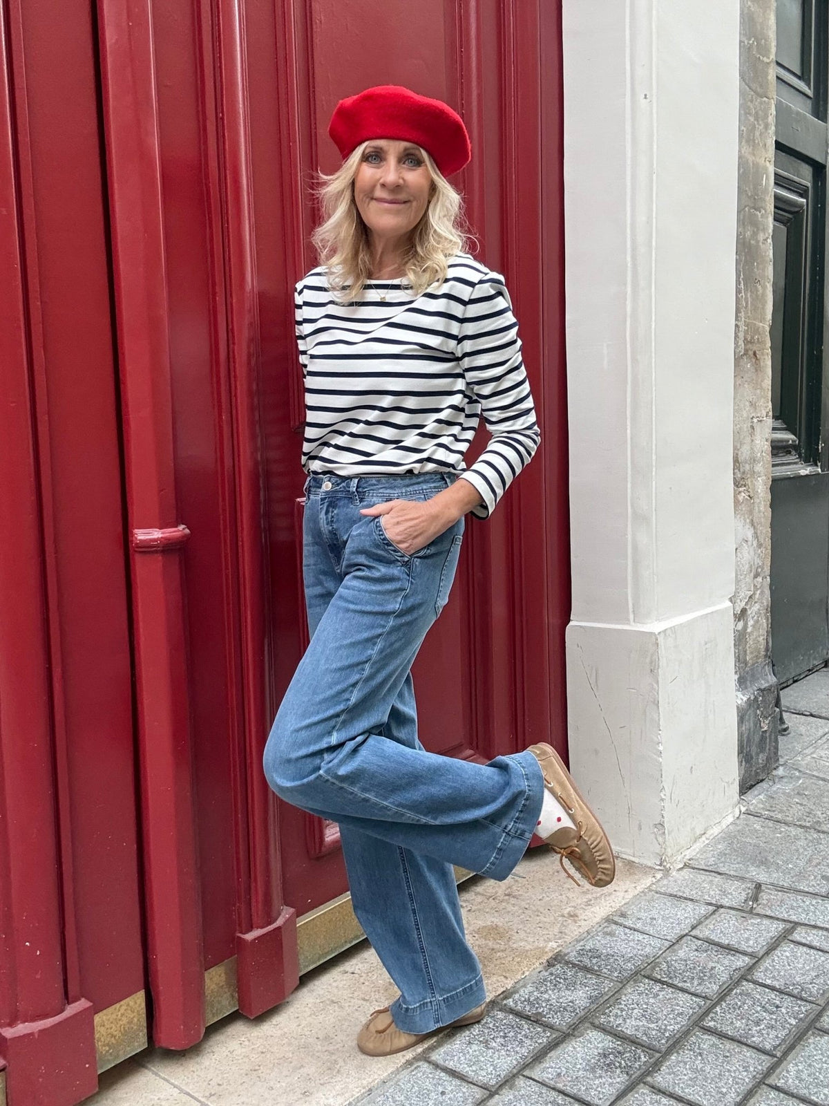 En kvinde med rød baret, stribet skjorte og Red Button Colette L.31-jeans står ved en rød dør på et fortov i byen.