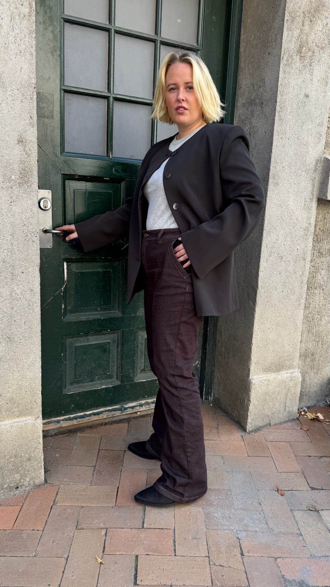 En person med kort blondt hår, iført en sort blazer og en rød Button Bibette Mørkebrun, står ved en grøn dør på en murstensvæg.