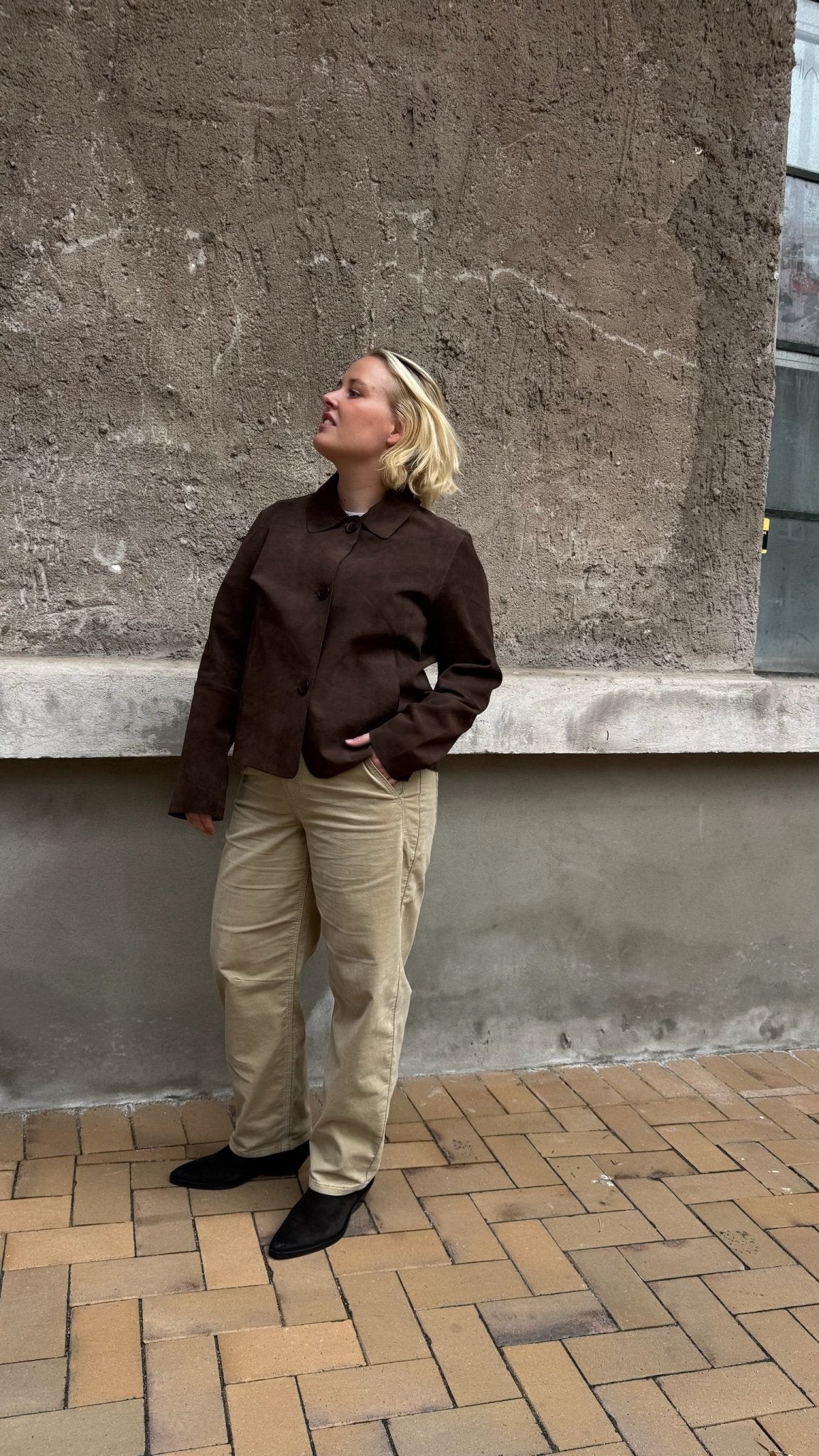 En person iført en brun jakke, beige bukser fra Red Button Caitlina og sorte sko står på en murstensgang ved siden af en væg med struktur.
