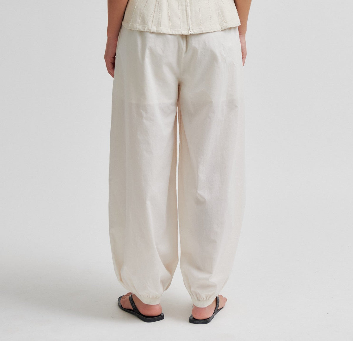 Person i 154638 | ANDEN KVINDE – Avida-bukser i offwhite og sorte sandaler, set bagfra på en lys baggrund.