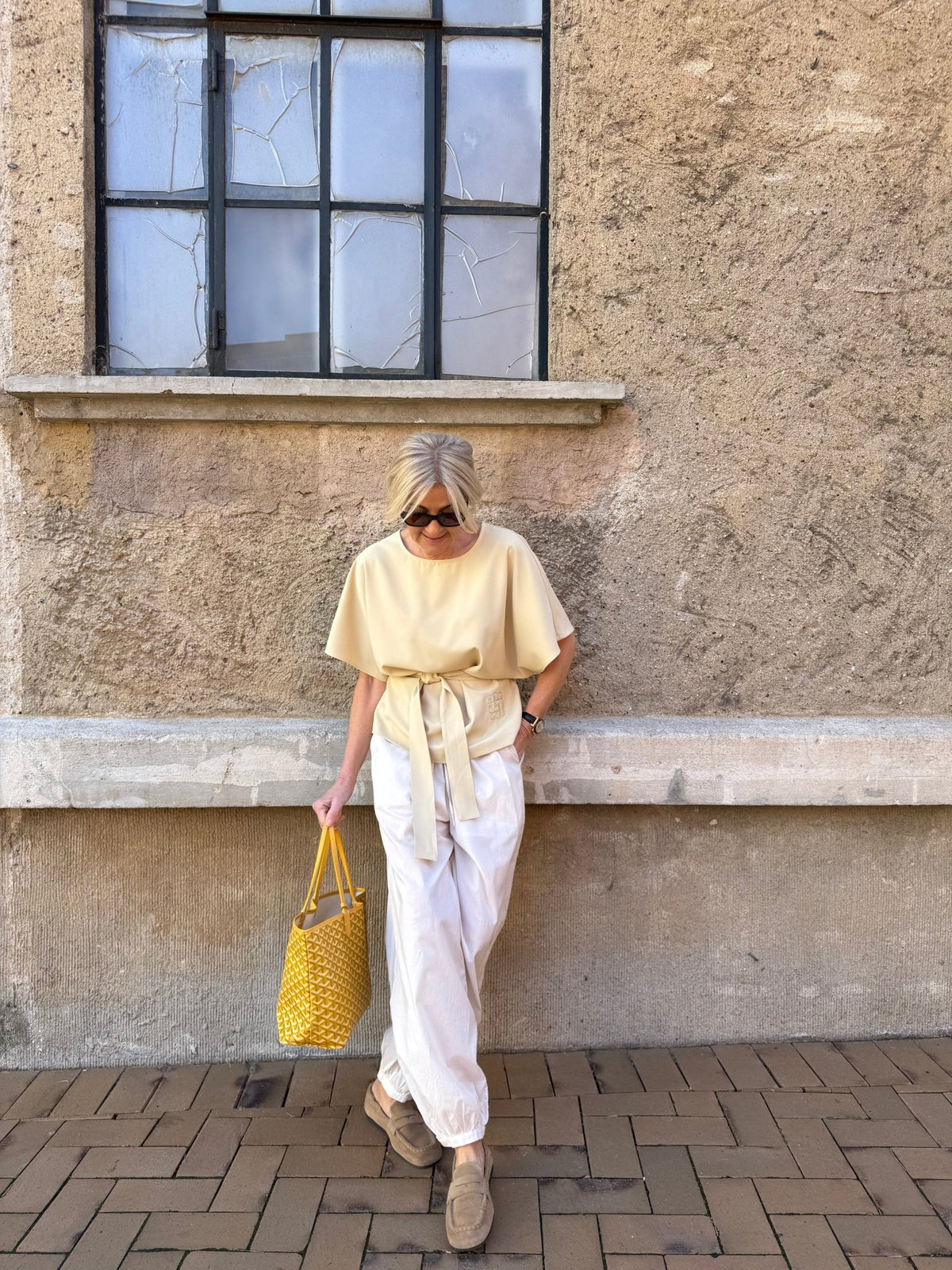 En kvinde læner sig op ad en stenmur iført en Avida-top i farven »SECOND FEMALE Avida Off white«, løse bukser, loafers og en gul totebag.
