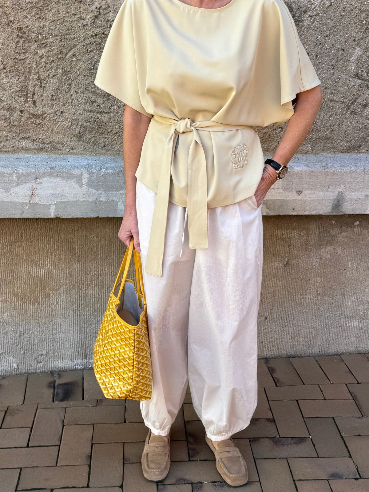 En person i en lysbeige bluse, ANDEN KVINDE: Avida-bukser i offwhite, lysebrune sko og en gul taske ved en stenmur.