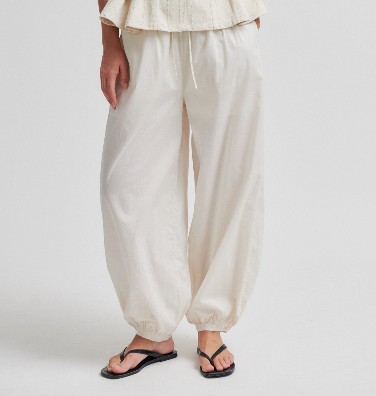 Model iført SECOND FEMALE Avida-bukser i offwhite med elastiske manchetter og sandaler, fotograferet på en ensfarvet baggrund.