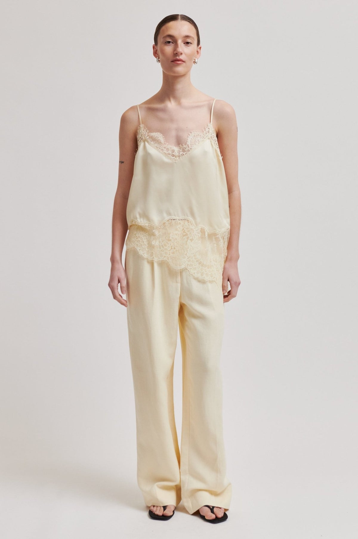 Modellen bærer en cremefarvet blonder-camisole og bukser med brede ben fra SECOND FEMALE Wilina Lysegul foran en hvid baggrund.