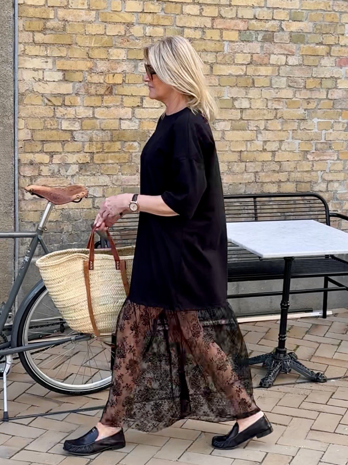 En blond kvinde i en sort kjole fra VILA Vikata går forbi en cykel, mens hun holder en stråtaske i hånden ved en mur og et udendørsbord.