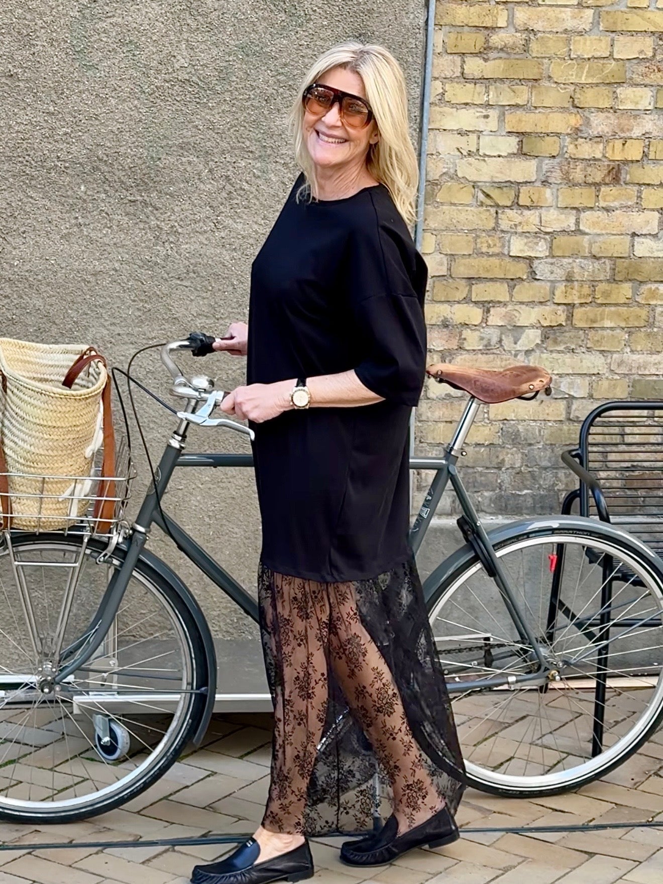 En smilende kvinde med solbriller står ved siden af en cykel iført VILAs Vikata Sort-kjole og sorte sko.