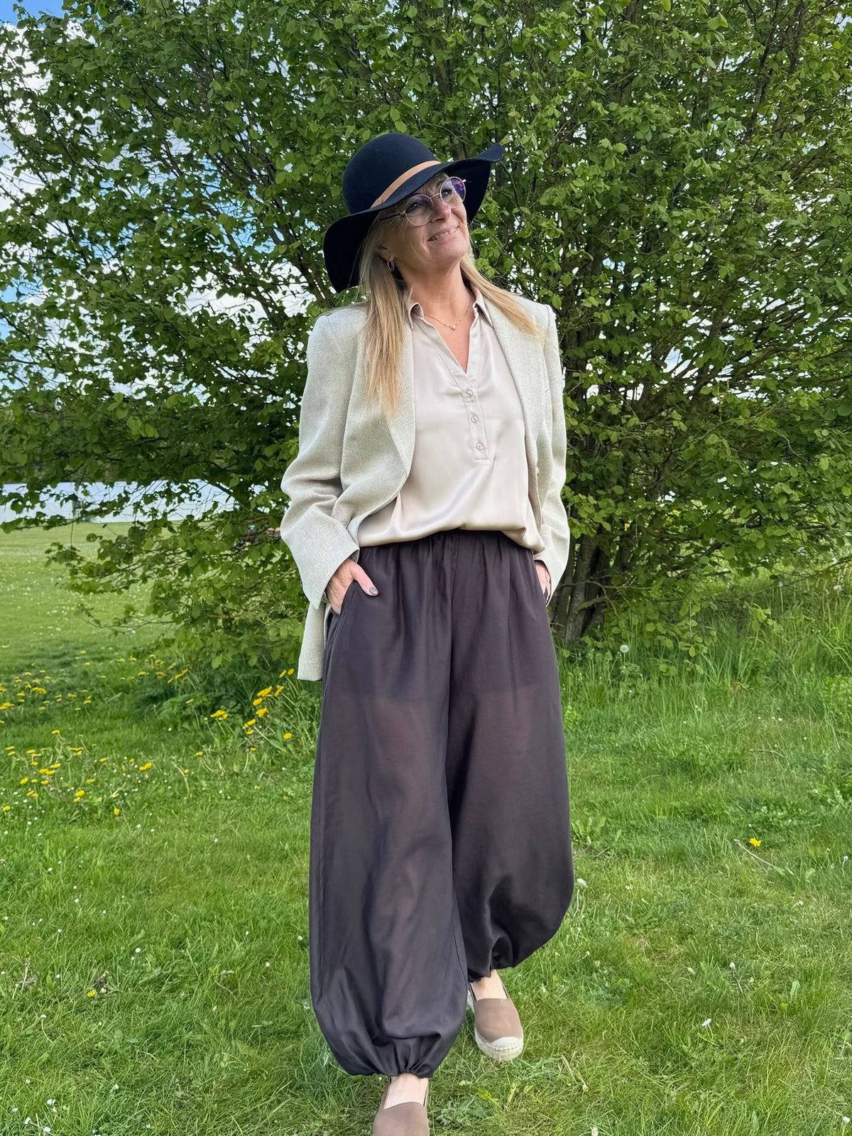 Personen bærer en hat, briller, en blazer, løse mørke bukser og en VILA - Vienna Ravenna Beige-bluse og står foran et løvrigt træ.