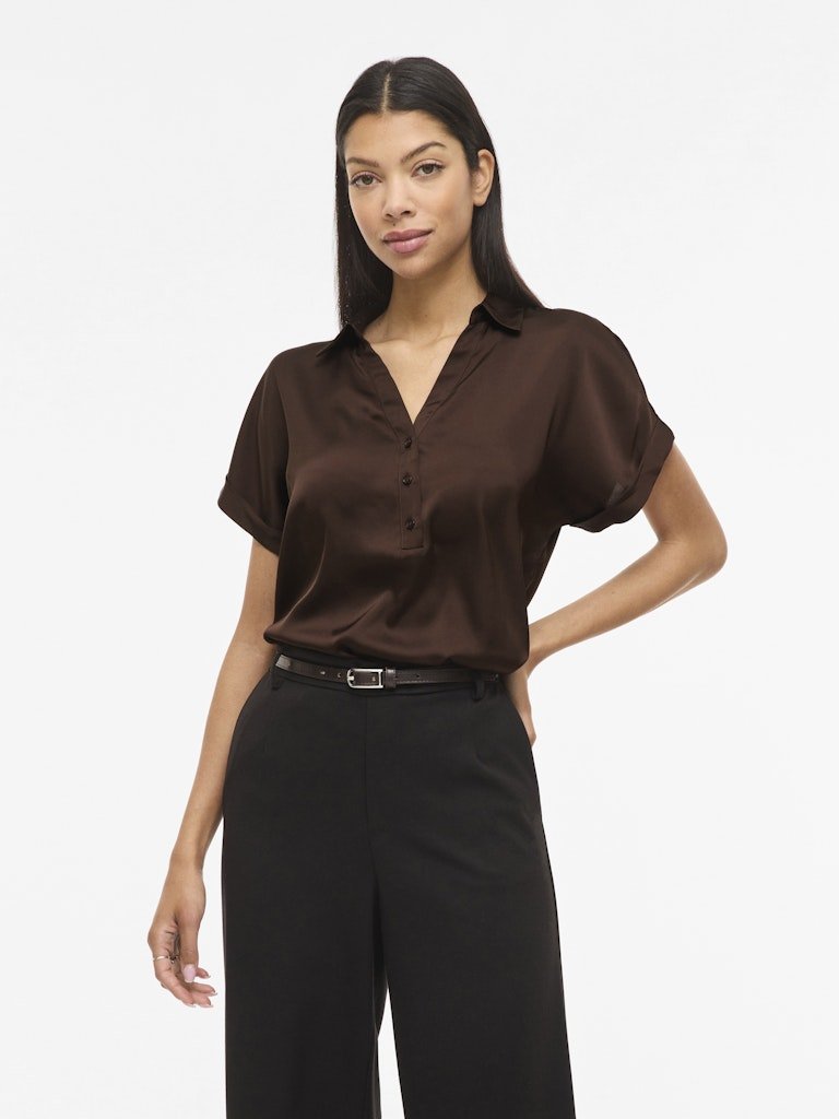 En kvinde bærer en VILA Vienna Ravenna Kaffe-bluse, 100  polyester, sammen med sorte bukser, foran en ensfarvet hvid baggrund.