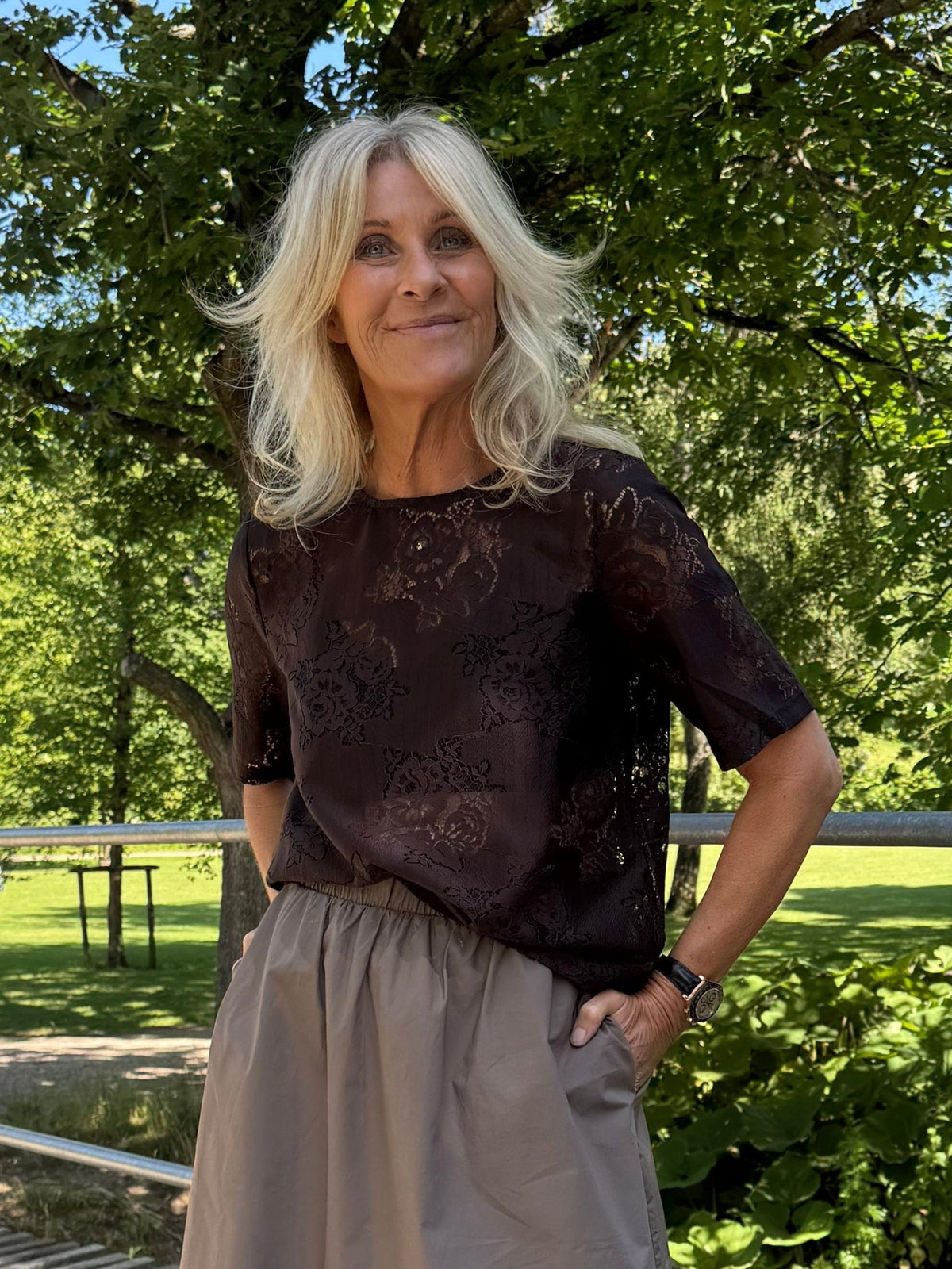 En blond kvinde står udenfor ved nogle grønne træer, iført en VILA Vimaven Mørkebrun bluse og et gråt nederdel.