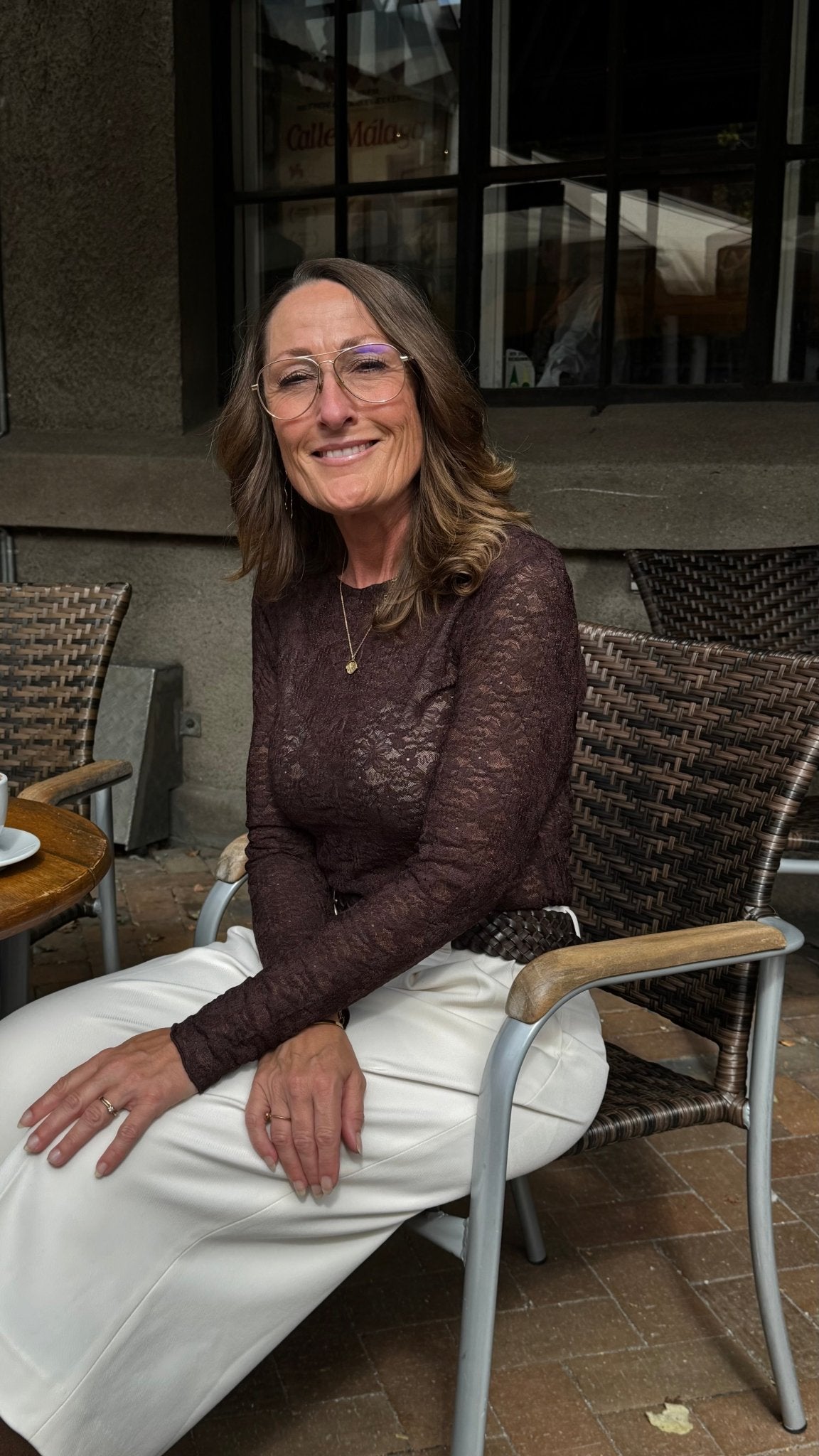 En kvinde iført en VILA Fay Kaffe-top og hvide bukser smiler ved et bord udenfor på en Coffee Bean-café.