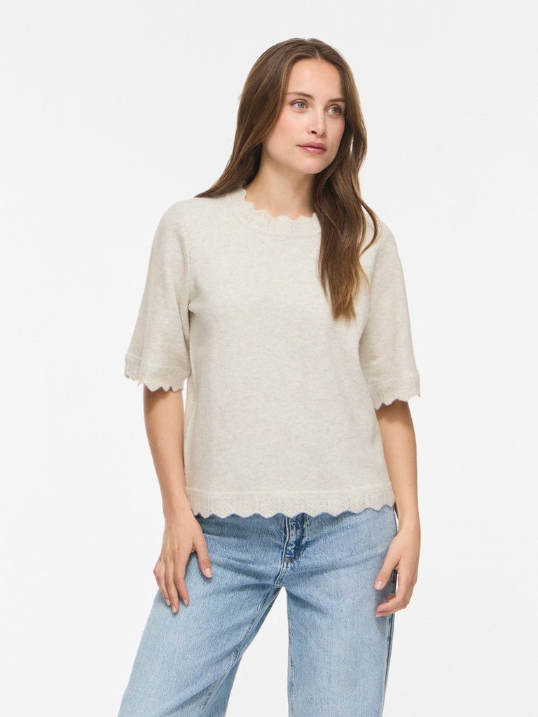 En kvinde med langt brunt hår har en VILA Vinikolina Lysegrå strikbluse og blå jeans på, der står i kontrast til det hvide.