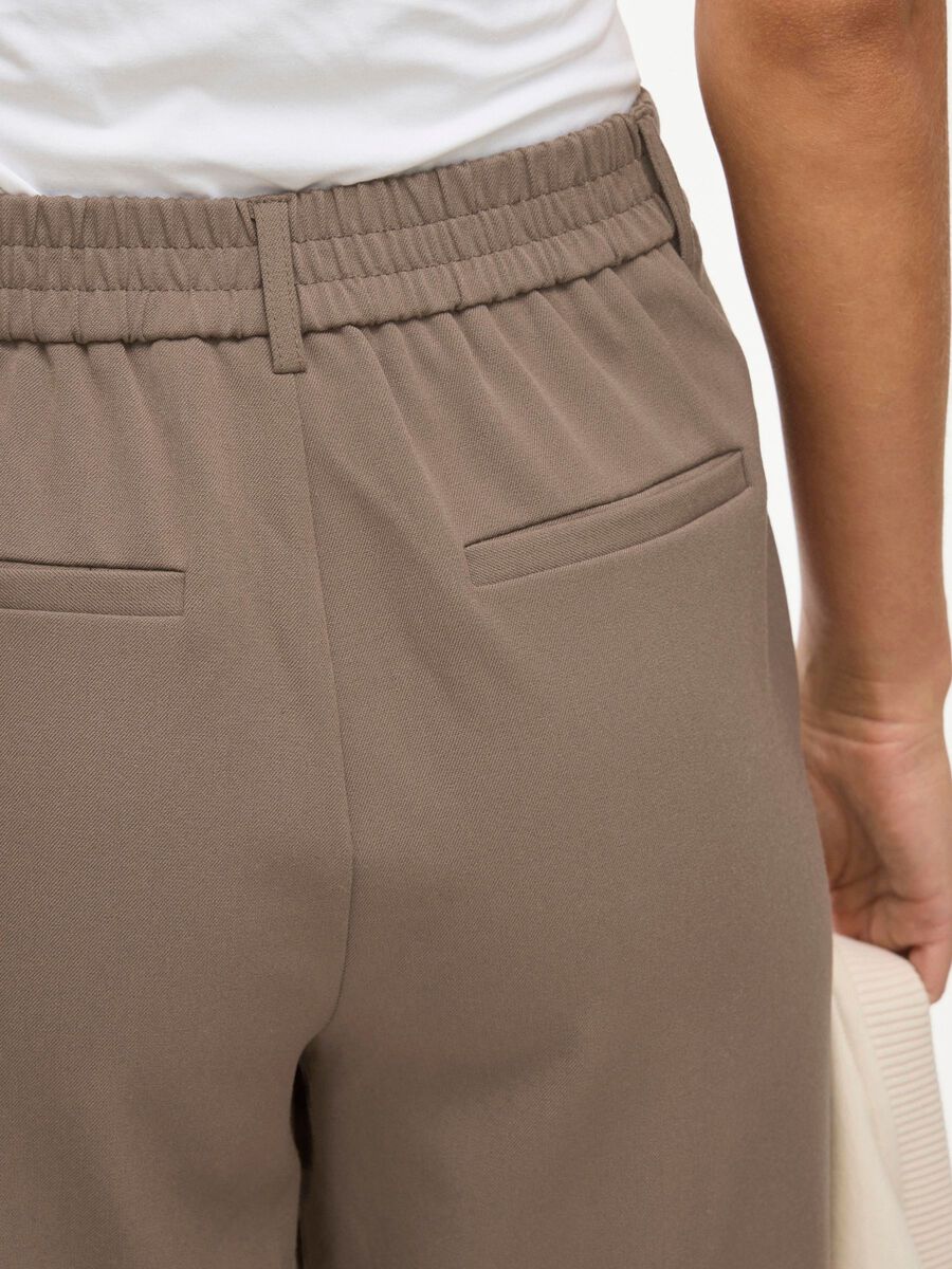 Nærbillede af en person, der har VILA - Vivarone Lysebrun-bukser på med elastisk talje og baglommer med kant, og som holder en beige genstand.