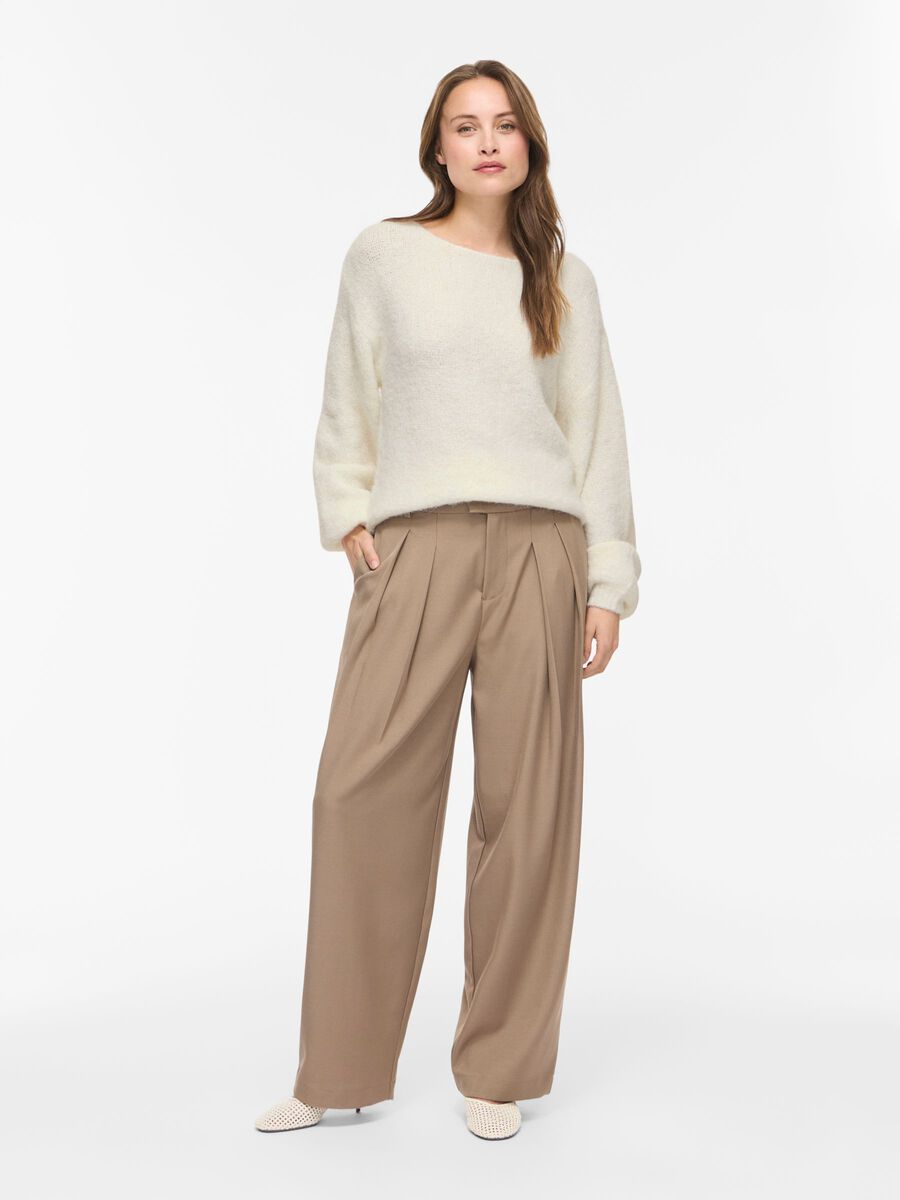 En kvinde viser VILAs Vimarley-top i offwhite frem sammen med beige bukser med brede ben og hvide sko, mens hun poserer foran en hvid baggrund.
