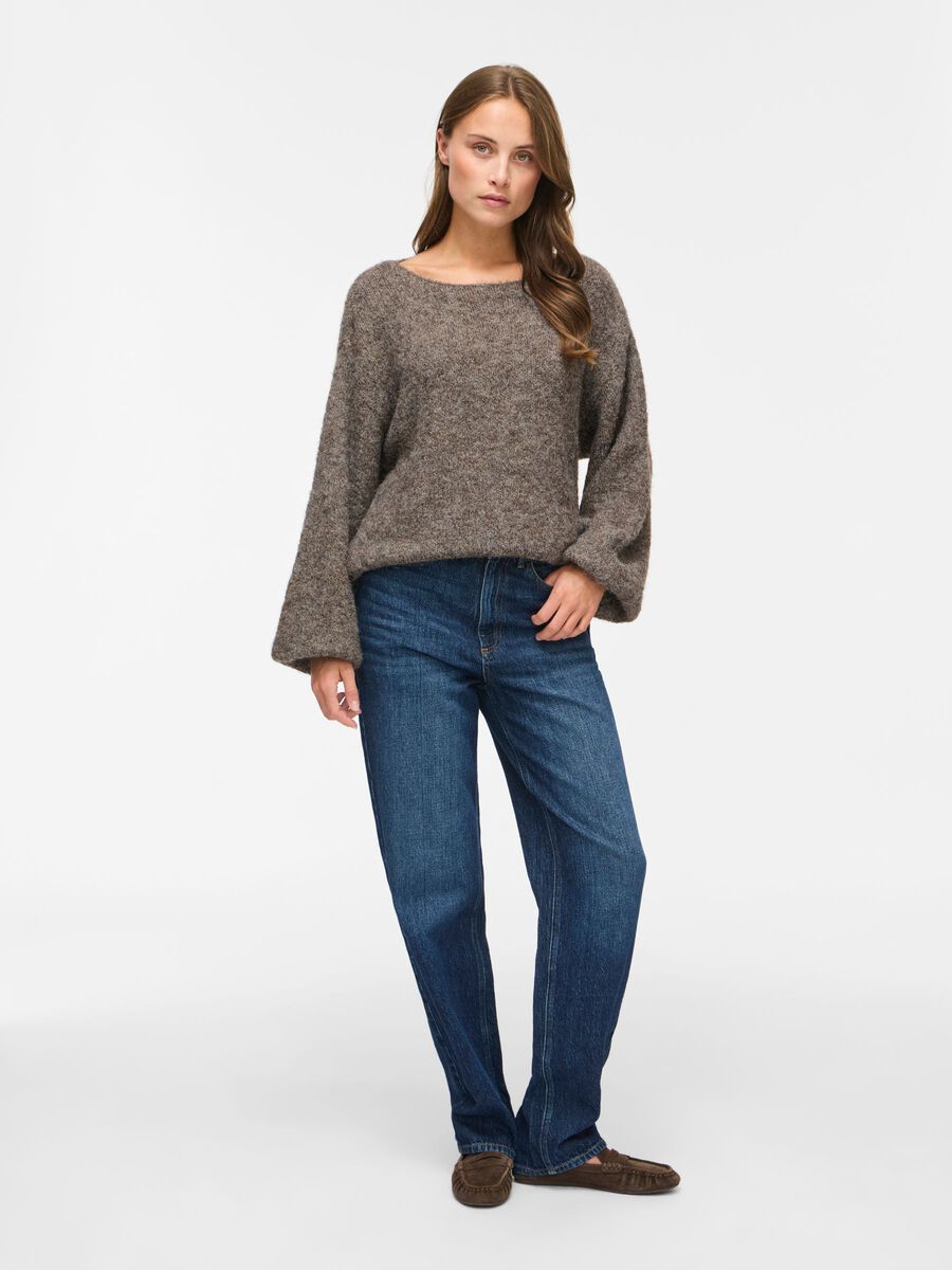 En kvinde er iført VILA – Vimarley Brunmeleret, blå jeans og brune sko og står foran en ensfarvet hvid baggrund.
