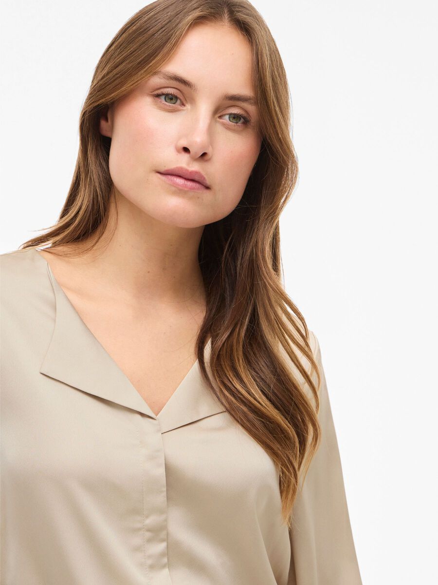 Kvinde med langt brunt hår iført VILA Viellette Beige 155327, der kigger til siden foran en ensfarvet baggrund.
