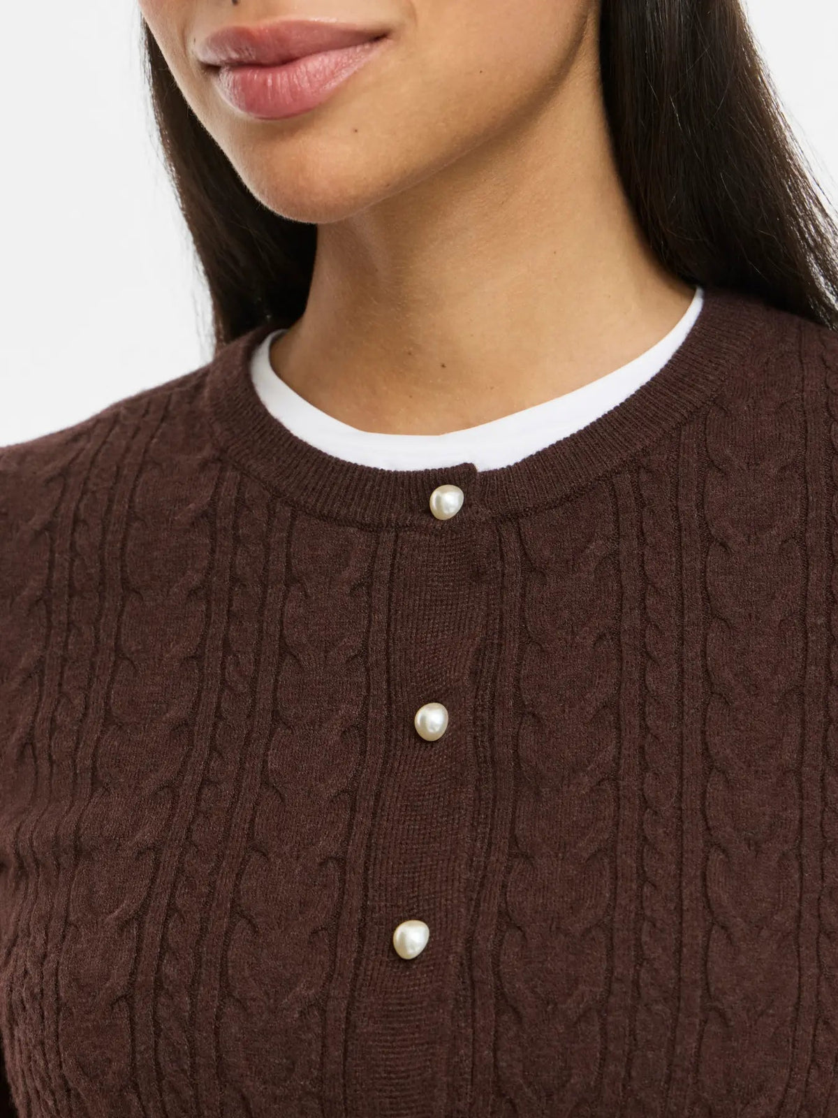 Kvinde iført en VILA Kerry Mørkebrun-cardigan med perleknapper over en hvid T-shirt med rund hals.