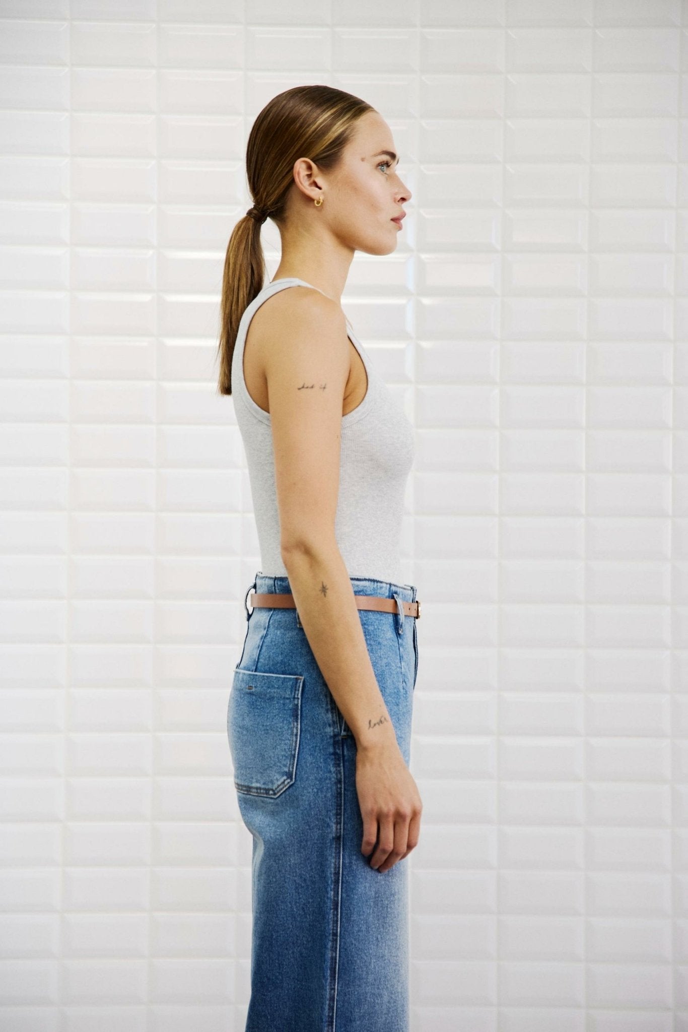 En kvinde med hestehale er iført en WithBlack 153548 Dordi Lysegrå ribbluse og blå jeans, set i profil foran en hvid flisebelagt væg.