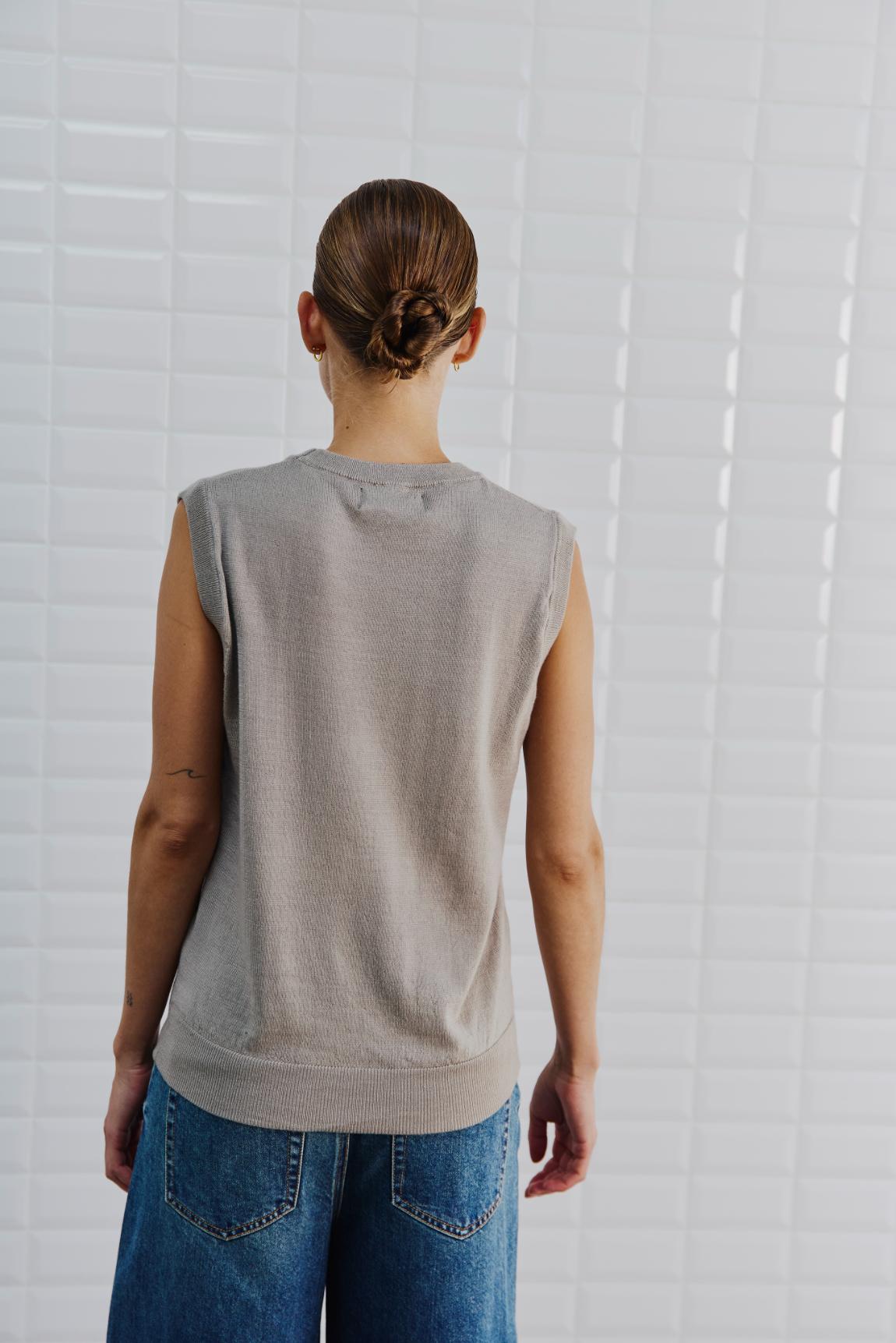 En person med en knold i håret, iført en WithBlack Miles Sand-vest og blå jeans, står med ansigtet vendt mod en hvid flisebelagt væg.