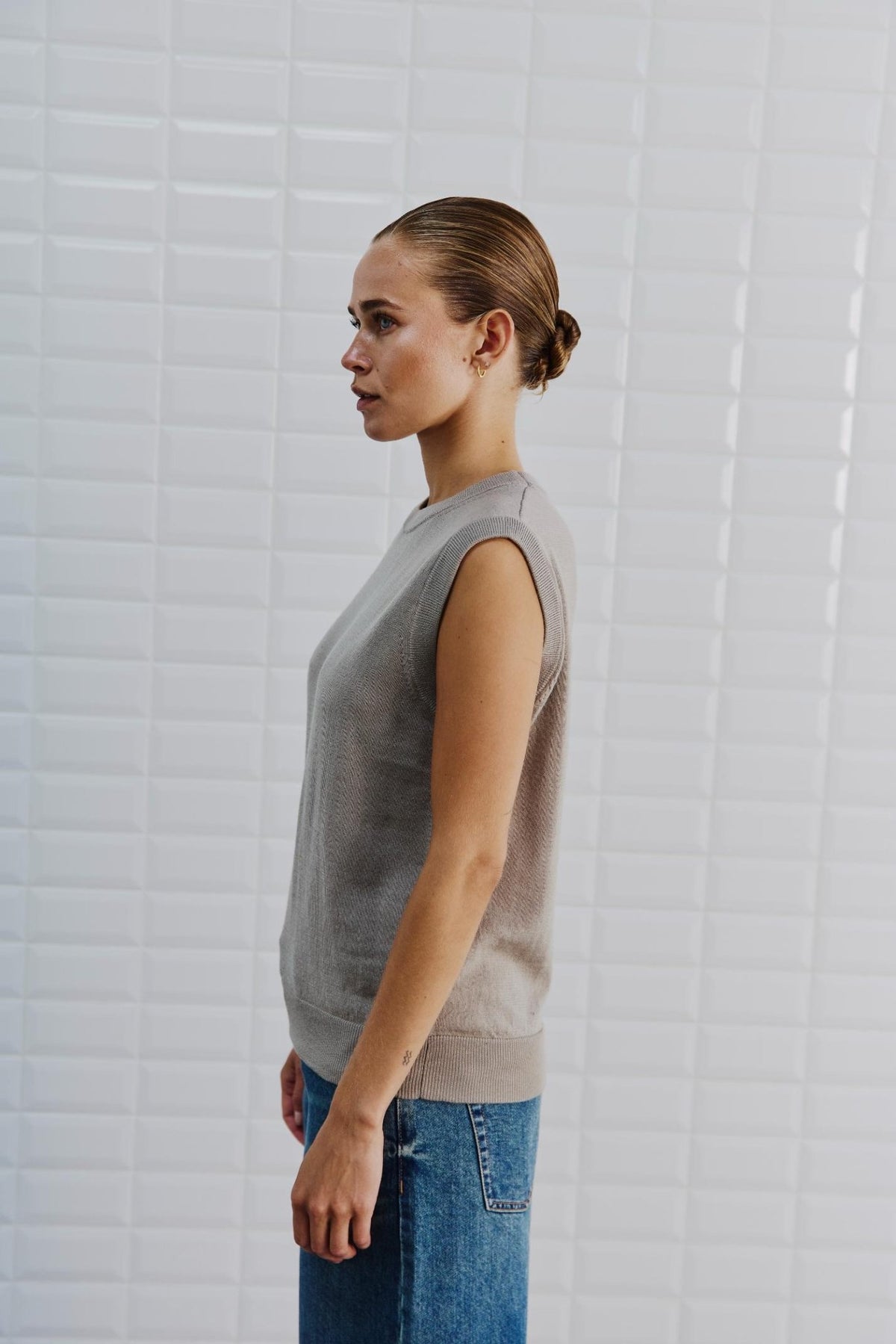 En kvinde iført en WithBlack - Miles Sand-vest og blå jeans står i profil foran en hvid flisebelagt væg.