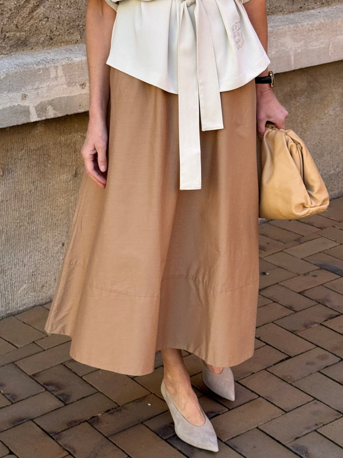 En person iført et WithBlack Milan Caramel-nederdel, en cremefarvet bluse med sløjfe, taupefarvede højhælede sko og en beige clutch på en murstensfortov.