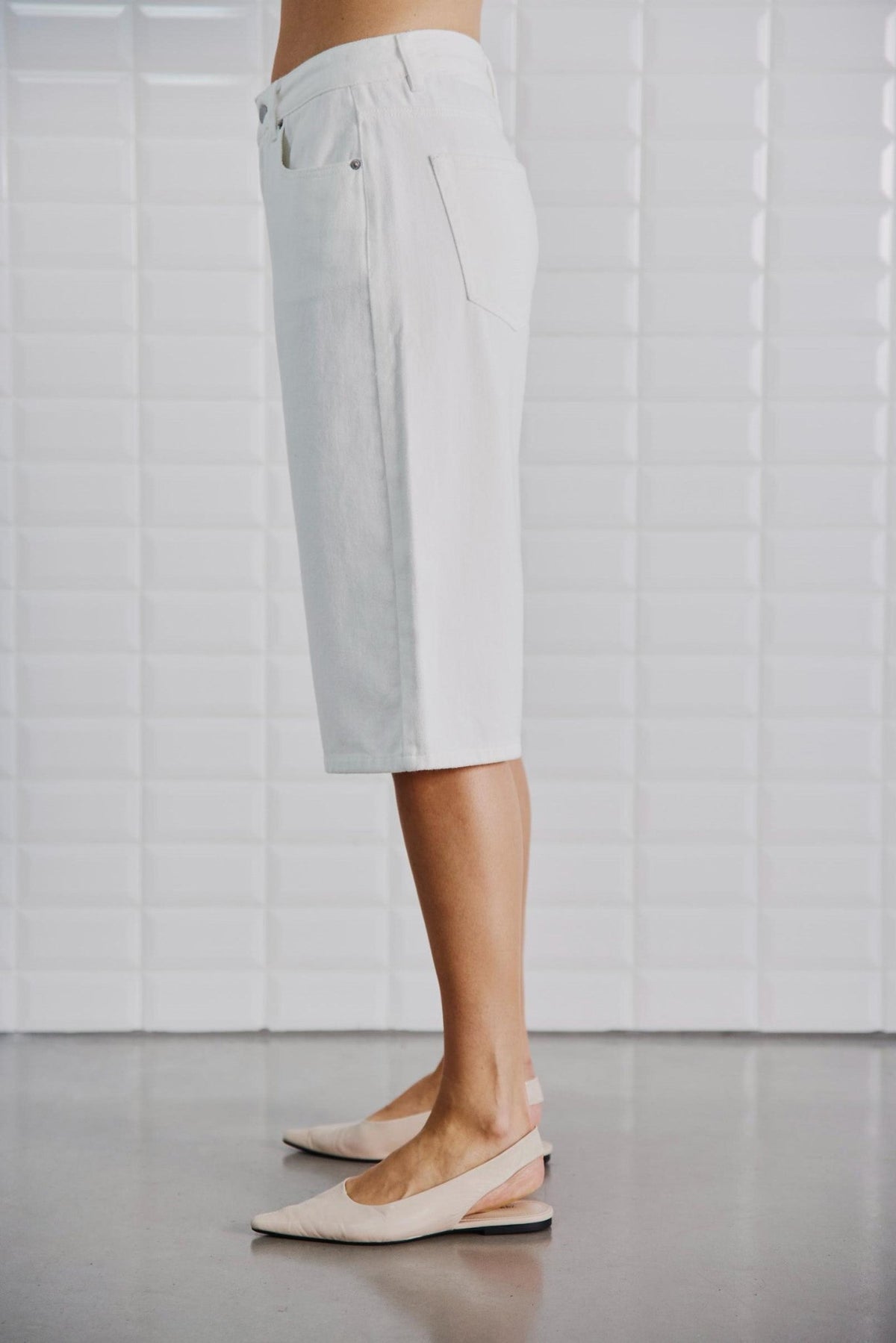 Modellen bærer et par off-white denimshorts fra WithBlack Gala og står med siden til en hvid flisebelagt væg, iført beige ballerinasko.