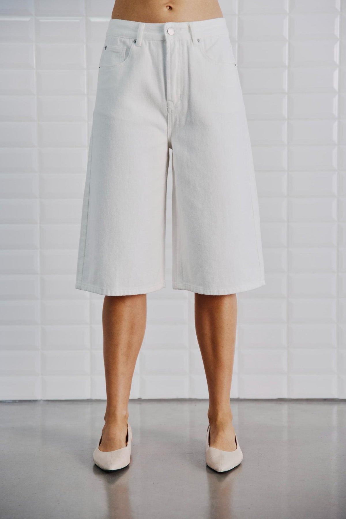 En person iført WithBlack – Gala Off White-culottes og beige flade sko, der står ved en hvid flisebelagt væg.