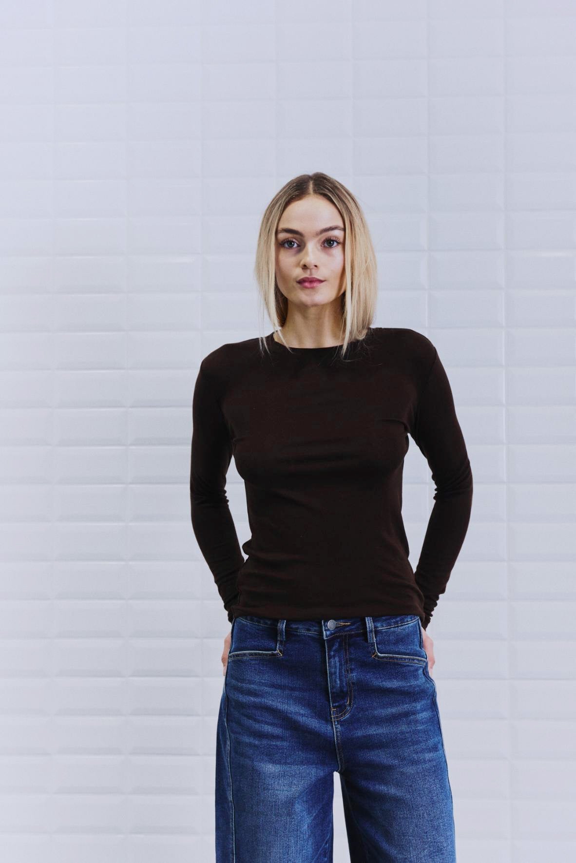 En kvinde iført en WithBlack Josefine Mørkebrun-bluse og jeans står foran en hvid, struktureret væg og ser direkte ind i kameraet.