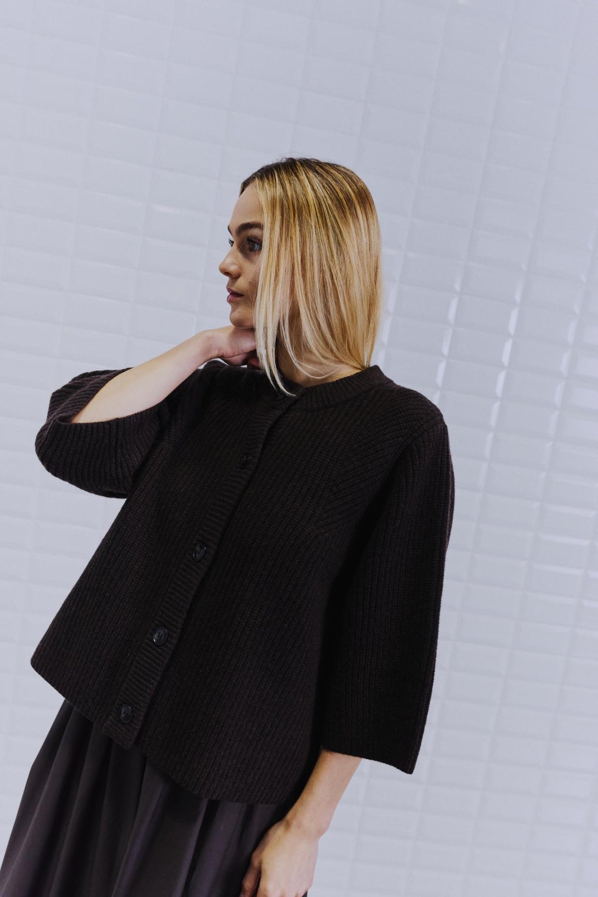 En kvinde med glat blondt hår iført en WithBlack Vivianna Mørkebrun-cardigan, der står ved en hvid flisebelagt væg.