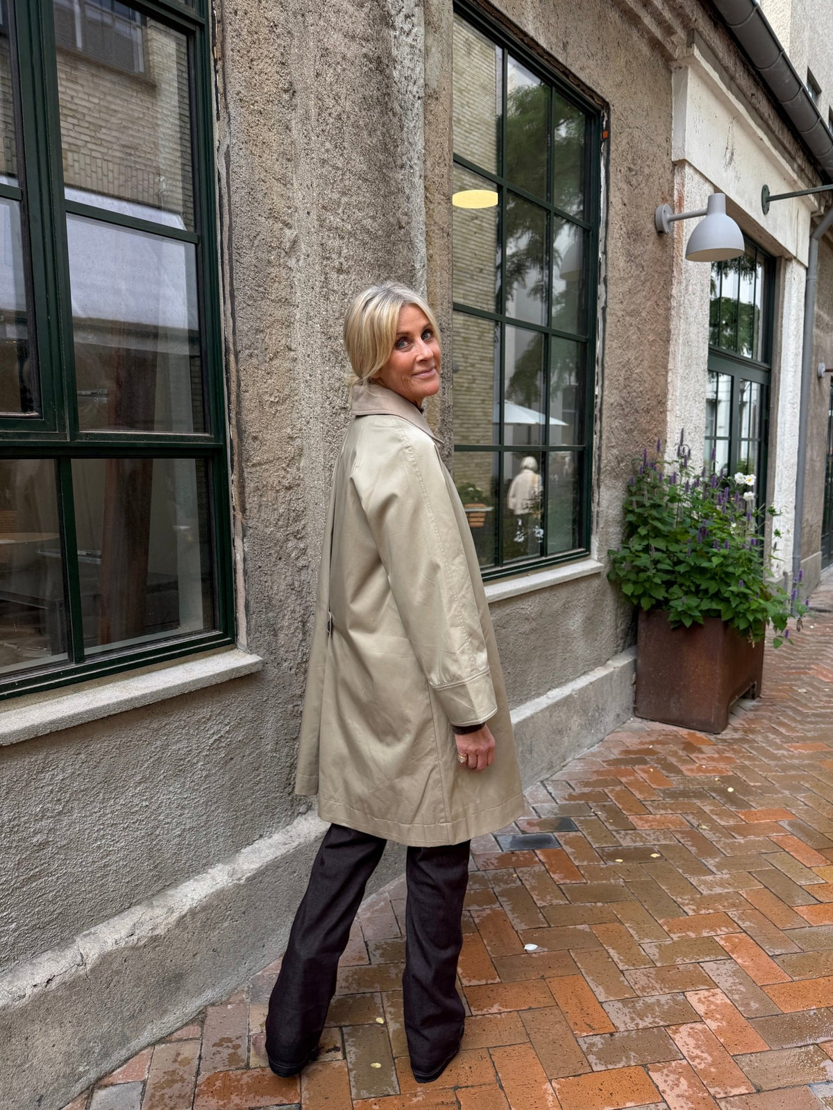 En person iført en ALAIA Sand-jakke fra WithBlack og mørke bukser står på en murstenssti ved siden af en bygning med store vinduer.