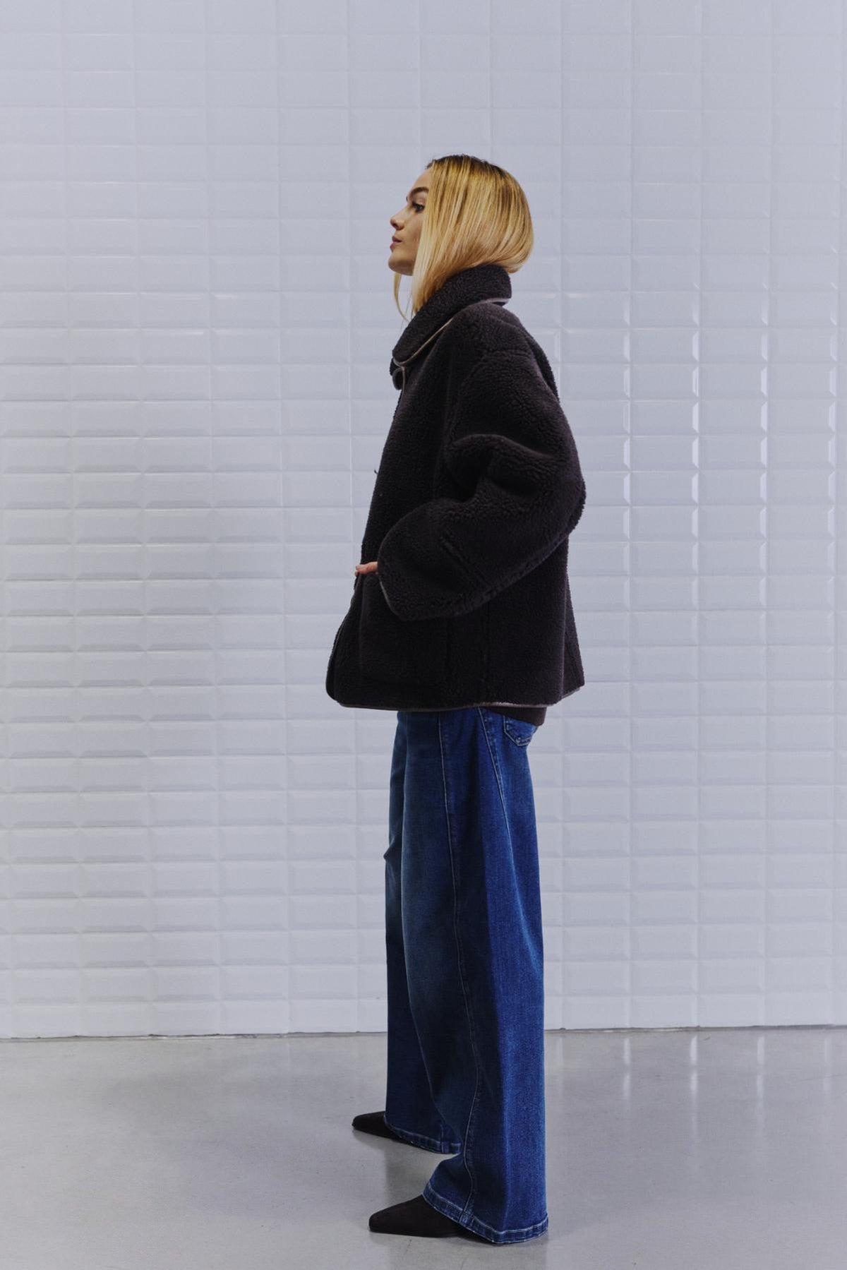 En kvinde med glat, blondt hår har en WithBlack IRIS Mørkebrun-jakke og jeans med brede ben på og står ved en hvid væg.