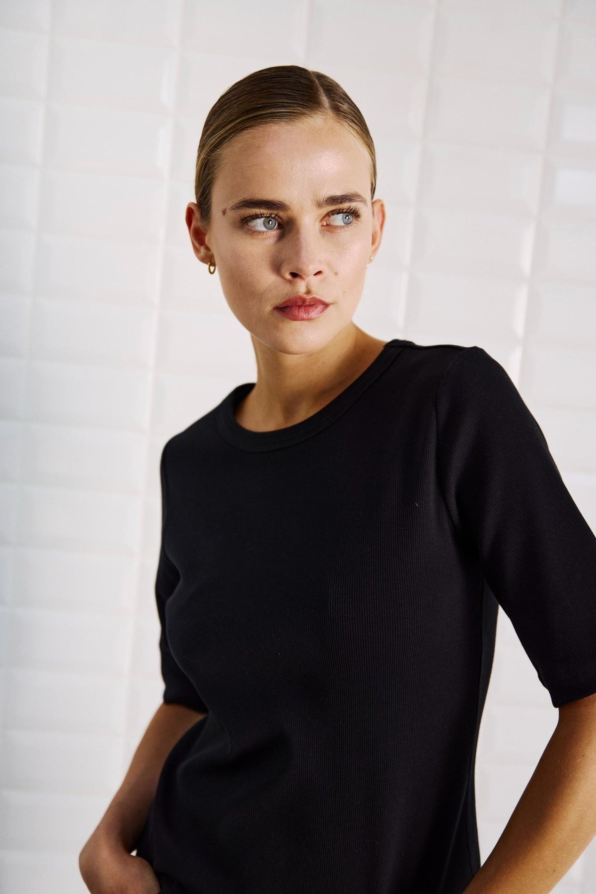 En model iført en Lullu Sort-T-shirt fra WithBlack står ved en hvidflisebelagt væg med håret glattet tilbage og blikket rettet mod siden.