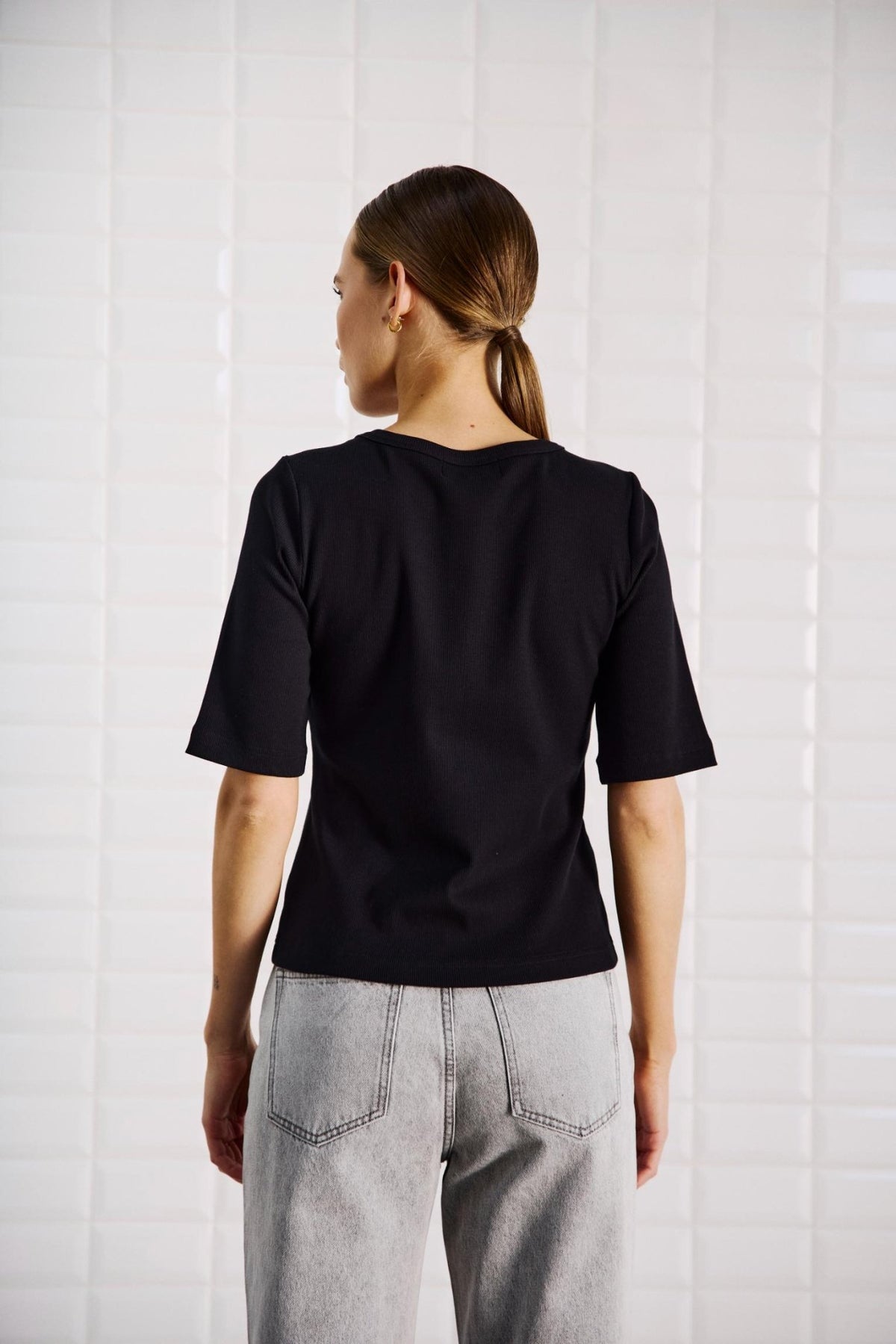 En person med glat, brunt hår, iført en WithBlack Lullu Sort-T-shirt og lyse jeans, set bagfra.