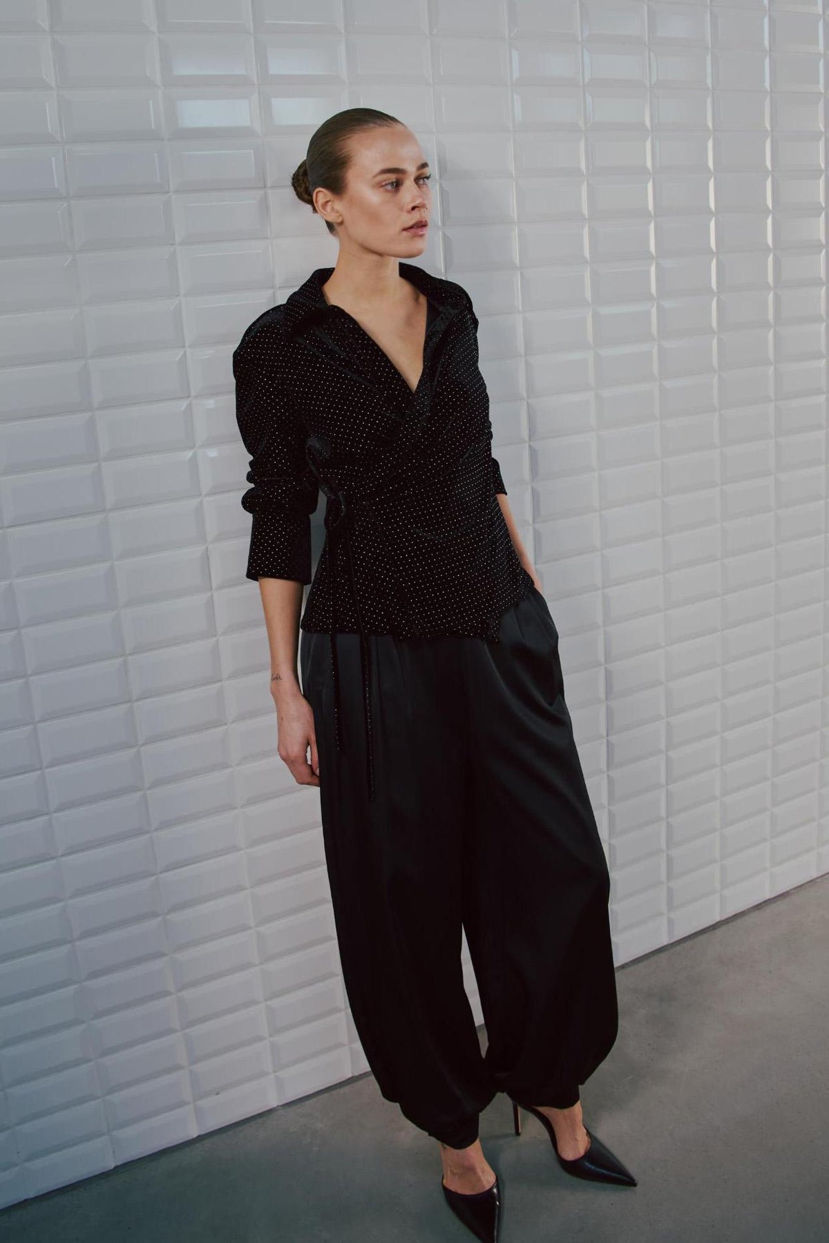 En kvinde iført model 156397 | WithBlack – Cyrus Sort: en sort langærmet wrap-top med prikker og satinbukser med brede ben.