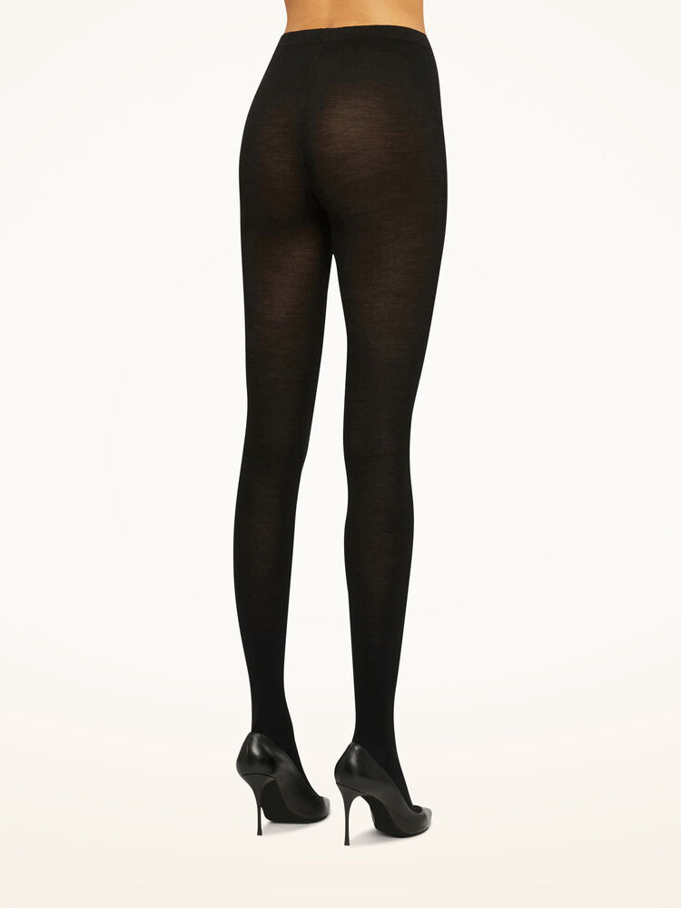 Model iført Wolford 035077 Merino Black W-strømpebukser og sorte højhælede sko, set bagfra på en lys baggrund.