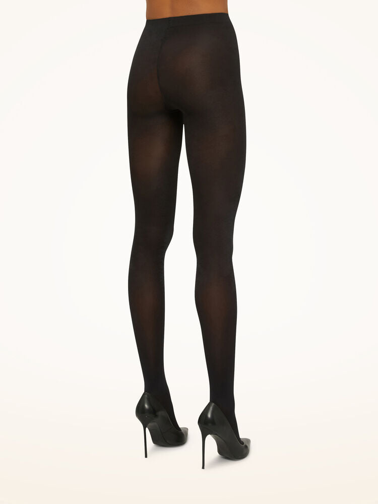 Model iført Wolford Satin Opaque 50 Black W-strømpebukser og sorte højhælede sko, set bagfra foran en ensfarvet lys baggrund.
