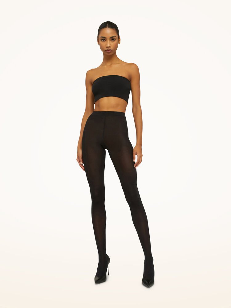 En kvinde iført Wolford 041567 Satin Opaque 50 sorte strømpebukser og en sort tube-top står foran en ensfarvet lys baggrund.