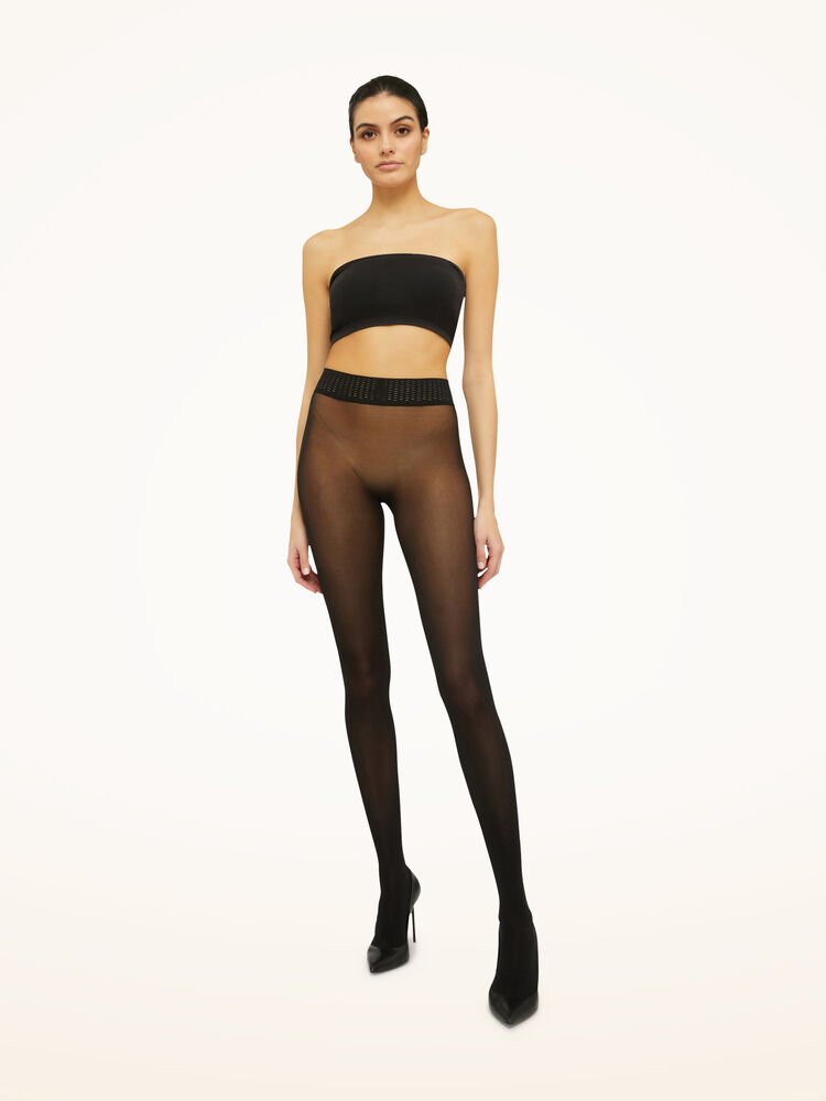 En kvinde iført en sort bandeau-top, Wolford Fatal 50 Seamless Black W-strømpebukser og højhælede sko står foran en ensfarvet baggrund.