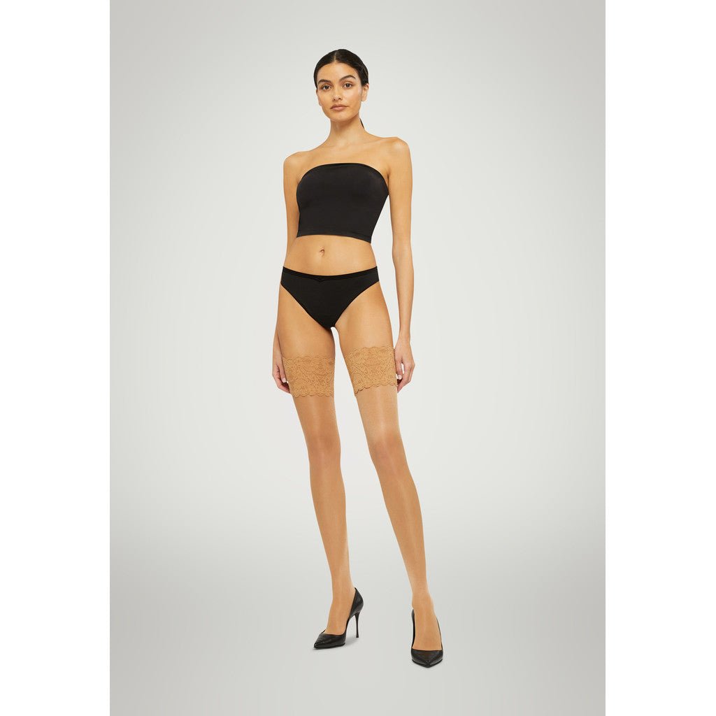 En kvinde iført en sort bandeau-top, Wolford Satin Touch 20 Gobi-strømper og højhælede sko står foran en ensfarvet baggrund.