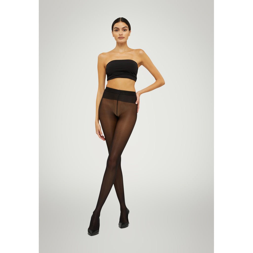 En kvinde i en sort bandeau-top og Wolford Neon 40 Black W-strømpebukser poserer foran en lys baggrund.